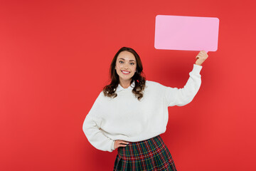 Wall Mural - positive brunette woman in sweater holding empty speech bubble on red background
