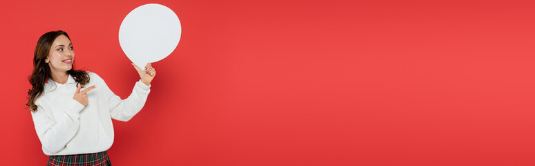Wall Mural - young woman in warm sweater pointing with speech bubble with copy space on red background, banner
