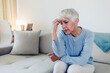 © Jelena Stanojkovic - Sad depressed woman at home sitting on the couch, looking down and touching her forehead, loneliness and pain concept. Depressed and sad grey hair mature woman sitting on bed at home during the day.