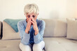 © Jelena Stanojkovic - Sad depressed woman at home sitting on the couch, looking down and touching her forehead, loneliness and pain concept. Depressed and sad grey hair mature woman sitting on bed at home during the day.