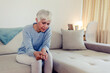 © Jelena Stanojkovic - Photo of elderly lady is enduring strong ache while sitting on the sofa during the day. Senior woman at home suffering with arthritis. Upset stressed mature middle-aged woman feeling pain ache.