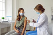 © Studio Romantic - Vaccination procedure. Smiling brave teenage girl wearing mask receives dose of coronavirus vaccine at vaccination center. Female doctor holds syringe and disinfects injection site on child's hand.