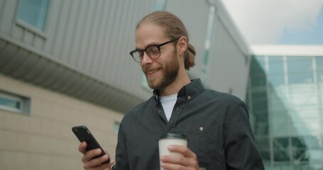 Man in eyeglasses uses mobile phone and holding cup of coffee during walk