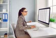 Busy Financial Accountant Working On Modern Desktop Computer. Serious Woman In Suit And Glasses Sitting At Office Desk, Working With Electronic Business Spreadsheet Files And Taking Notes In Her Book