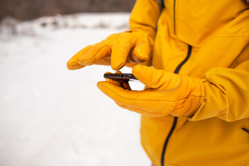 Wall Mural - Close-up of male hands in gloves hold the phone and dial the message. The man dresssed yellow warm jacket and trousers, bagpack traveling in the snow landscape