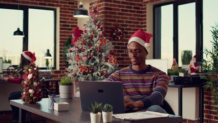 Wall Mural - African american man browsing internet on laptop and wearing santa hat to celebrate christmas eve in startup office. Male employee working on company report in festive space with winter decor.