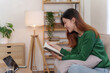 © amnaj - Happy beautiful Asian female college student sitting online studying and taking notes in a notebook to submit university work in the living room during the holiday.
