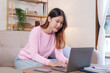 © amnaj - Bright, cute and happy Asian female student study online in the living room using laptop computer in a relaxed manner. Young Asian woman using laptop at home.