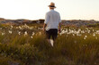 A caucasian man with straw hat and a white linnen shirt walking on a field at sunset.