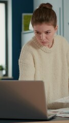 Wall Mural - Portrait of focused startup employee looking at laptop and charts comparing sales statistics standing at desk. Woman in busy office sorting through business papers looking at data analytics.