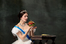 Wow. Young Beautiful Woman, Royal Person, Queen Or Princess In White Medieval Outfit Tasting Burger On Dark Background. Comparison Of Eras, Modern, Fashion, Beauty.