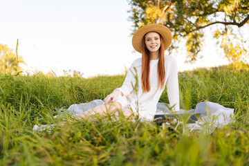 Wall Mural - portrait of smiling young red-haired young woman in straw hat and white dress sitting posing with la