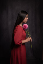 Young Asian Woman In Red Dress Holing Pink Dahlia Flower In Dark Painterly Studio Setting