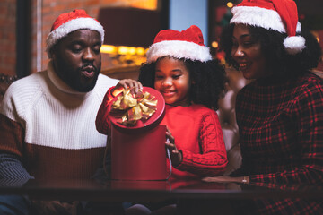 African American families happy and laughing celebrate Christmas eve sitting in leaving room decorations with a green Christmas tree and lighting, giving a surprise red gift box.