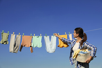 Wall Mural - Smiling woman hanging baby clothes with clothespins on washing line for drying against blue sky outdoors