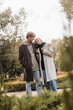 © LIGHTFIELD STUDIOS - happy and young man in coat hugging blonde girlfriend while standing around plants in park.