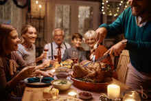 Close Up Of Man Carving Turkey Meat During Thanksgiving Meal With His Extended Family.