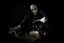 An Evil Cursed Nun With A Notebook And A Cross In Her Hands Looks At The Camera On A Black Background. Halloween. Horrors.
