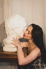 A woman with black hair and bright make-up touches the head statue with her face and hands. Has red lips and beautiful eyelashes. Long black hair falls on the back. Dressed in a corset. On a white bac