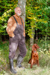 Casually in a country style with dungarees, sweater, rubber boots, an elderly man leans against a tree trunk and enjoys the colorful nature in autumn together with his Irish Setter hunting dog.
