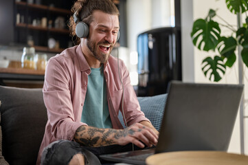 Young cheerful man with a sleeve tattoo using a laptop computer and listening to music online over headphones while working