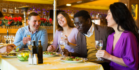 Wall Mural - Cheerful adult people of different nationalities enjoying evening meal in restaurant. Friendly meeting over dinner with wine ..