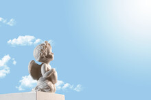 Figurine Of An Angel Cupid On The Podium With A Heart On A Blu Sky With Clouds . Valentine's Day.