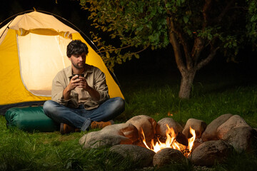 male tourist settles down next to a tent by the campfire and drinks coffee or tea from a thermos in 
