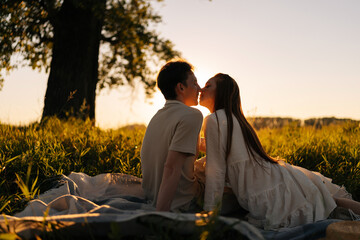 Wall Mural - silhouette of happy young couple sitting hugging and kissing on green field having picnic date in fi