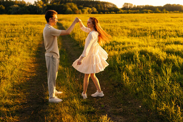 Beautiful young woman in dress dancing with loving boyfriend on green meadow in summer evening during sunset with soft sunlight. Happy relationships between couple enjoying spending time together.