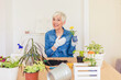 © Jelena Stanojkovic - Smiling senior woman cultivating potted plants at home. Relaxing hobby. Caucasian woman gardener in casual clothes and protective gloves taking care of house plants, concept of home garden and hobby.