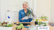 © Jelena Stanojkovic - Lovely senior housewife with flower in pot and gardening set indoors. Portrait of elderly woman gardening at home. Retired female care for her plants.