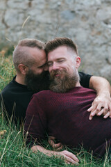 Wall Mural - Hipster fashionable  gay couple having fun in grass.  Long beard male homosexual partners lying together outside, holding hands, enjoying in romance outdoors with smiles on faces.