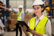 Happy female warehouse engineer with safety vest using tablet for checking goods and supplies on shelves with goods background in warehouse store, Logistic and business export concept