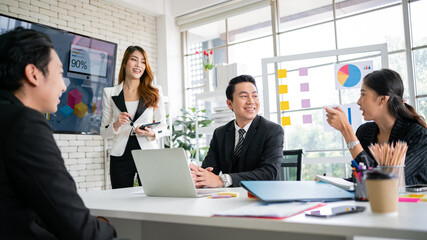 A cheerful and confident Asian businesswoman stands and uses a tablet to present a bar chart of data from a monitor to her office colleagues. Focus on Asian business women leader role at the meeting.