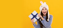 Happy Teen Girl Wear Earflap Hat Holding Purchase Box On Yellow Background. Kid Girl With Gift, Horizontal Poster. Banner Header With Copy Space.
