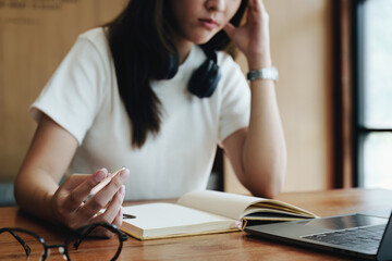 A portrait of a young Asian woman using a computer, wearing headphones and using a notebook to study online shows boredom and pain from video conferencing on a wooden desk in library