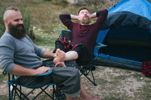 Hipster Long Beard Gay Couple Camping On Open Grass Field. Attractive Male Homosexuals Relaxing On Chairs In Front Of Tent In Late Autumn. Man Holding Partner's  Barefoot Legs In His Lap.