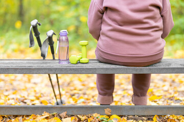 Rear view of fat elderly unrecognizable man resting sitting on a forest bench after playing sports. Scandinavian sticks, shaker, dumbbells. Modern lifestyle.