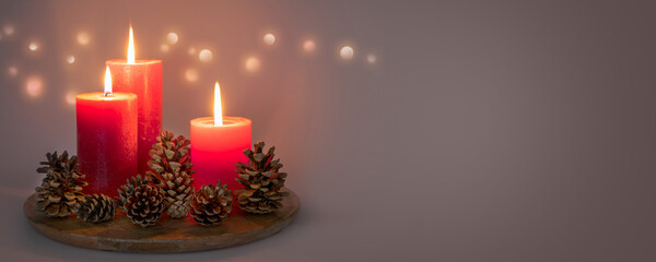 Wall Mural - three red burning candlelights with pine cones on grey background with copy space, bright illumination for christmas decoration in contemplative atmosphere at night