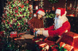 © ZaStock - Santa Clause packs gifts with his young girl helper while sitting at the table near fireplace.