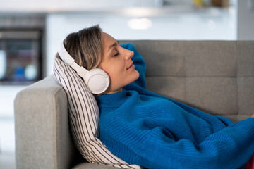 Relaxed blonde Caucasian woman with headphones sleeping lying down on comfortable sofa in living room. Casual girl with closed eyes listening to music or online radio with interesting podcasts