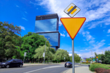 Give way priority sign and sign End Main road against the backdrop of blurred cityscape. Road signs at crossroads of European city. Priority signs in foreground. In background is skyscraper, cars