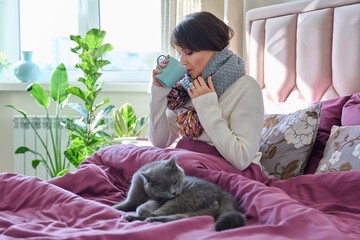 Wall Mural - Woman sitting at home in bed, warming with scarf and mug, with gray cat