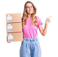 Poster - Young caucasian woman with blond hair holding delivery package smiling happy pointing with hand and finger to the side