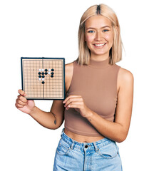 Sticker - Beautiful young blonde woman holding asian go game board smiling with a happy and cool smile on face. showing teeth.