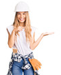 © Krakenimages.com - Beautiful caucasian woman with blonde hair wearing hardhat and painter clothes amazed and smiling to the camera while presenting with hand and pointing with finger.