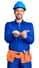 Canvas Print - Young handsome man wearing worker uniform and hardhat smiling with hands palms together receiving or giving gesture. hold and protection