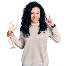 Young Hispanic Woman With Curly Hair Holding Small Wooden Manikin Smiling Amazed And Surprised And Pointing Up With Fingers And Raised Arms.