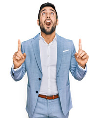 Wall Mural - Young hispanic man wearing business jacket amazed and surprised looking up and pointing with fingers and raised arms.